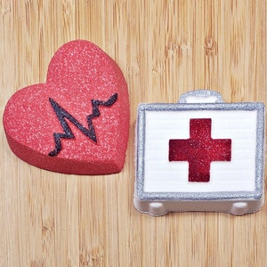 May include: A red heart-shaped bath bomb with a black EKG line design and a white first aid kit bath bomb with a red cross.