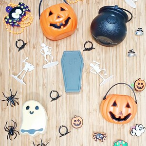 May include: A collection of Halloween-themed decorations on a wooden surface. The items include orange jack-o'-lantern buckets, a black cauldron, a gray coffin with "RIP" on it, spiders, skeletons, and ghost-shaped decorations.