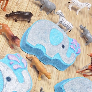 May include: Two blue and gray bath bombs shaped like elephants with pink water droplets. The bath bombs are surrounded by various animal figurines, including a giraffe, a tiger, a rhino, and a zebra.