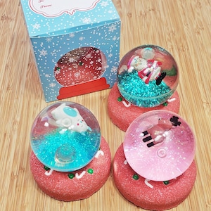 May include: Christmas-themed snow globes with glitter and miniature figurines, resting on red, gingerbread-shaped bases. A blue gift box with snowflakes and the text "Happy Holidays" is also present.