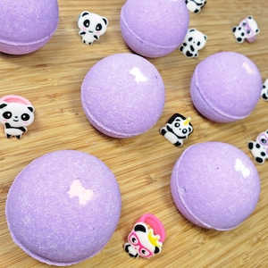 May include: Several round, lavender-colored bath bombs with a light pink bow design are scattered on a wooden surface. Small panda bear figurines in various poses are also present, adding a playful touch to the scene.