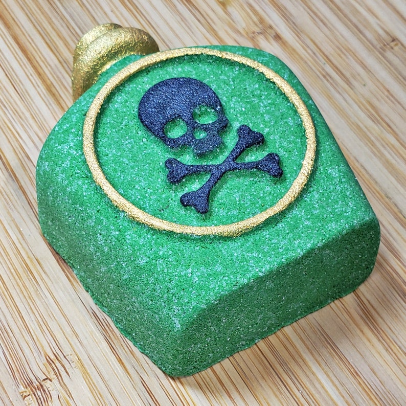 May include: A green, bottle-shaped bath bomb with a gold-colored top. The bath bomb has a black skull and crossbones design inside a gold circle. The bath bomb is on a wooden surface.
