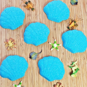 May include: Seven blue bath bombs shaped like seashells on a wooden surface with various sea-themed toys, including a starfish, a crab, a seahorse, and an octopus.