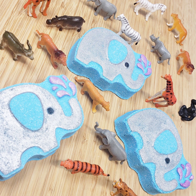 May include: Two blue and gray bath bombs shaped like elephants with pink water droplets. The bath bombs are surrounded by various animal figurines, including a giraffe, a tiger, a rhino, and a zebra.