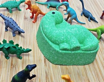 Peut inclure: Une bombe de bain en forme de dinosaure verte avec une surface texturée. La bombe de bain est entourée de divers jouets en plastique de dinosaures, dont un dinosaure vert et jaune, un dinosaure bleu, un dinosaure rouge et un dinosaure gris.