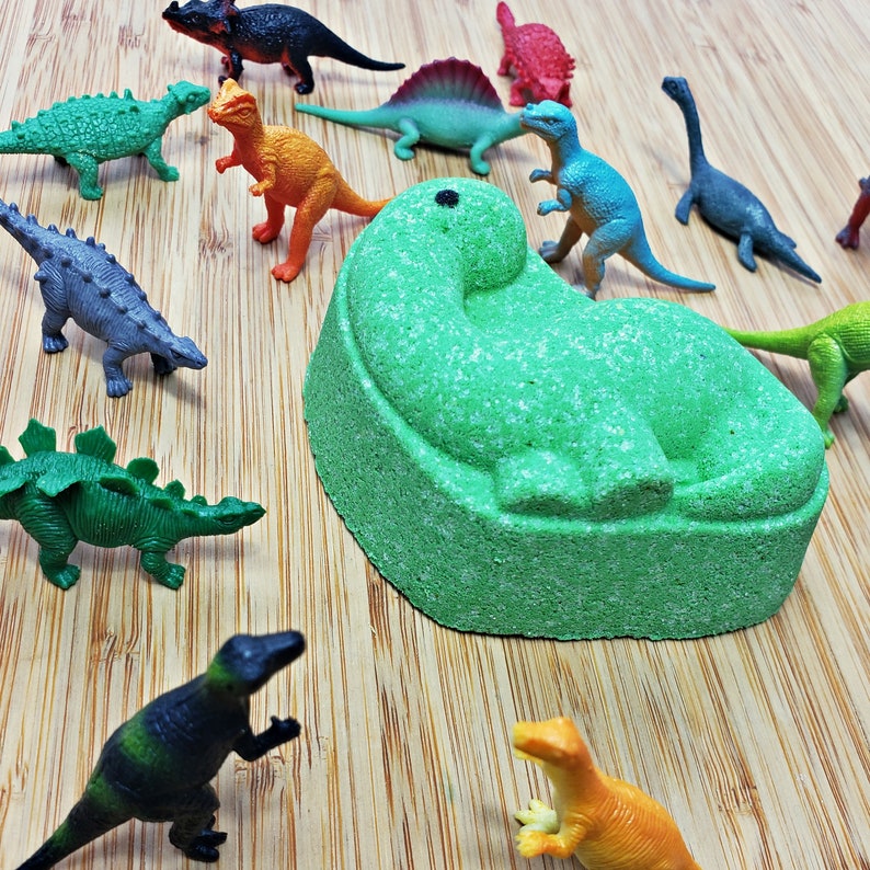 May include: A green dinosaur bath bomb with a textured surface. The bath bomb is surrounded by various plastic dinosaur toys, including a green and yellow dinosaur, a blue dinosaur, a red dinosaur, and a gray dinosaur.