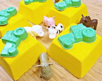 Peut inclure: Plusieurs bombes de bain jaunes en forme de cube, surmontées de tracteurs verts. Des figurines d'animaux de la ferme en plastique, dont un cochon, une vache, un mouton et un cheval, sont disposées autour. Fond en bois clair.