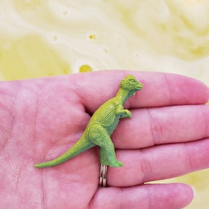 May include: A green plastic dinosaur toy in the palm of a hand. The dinosaur has a long tail and a small head with a crest.