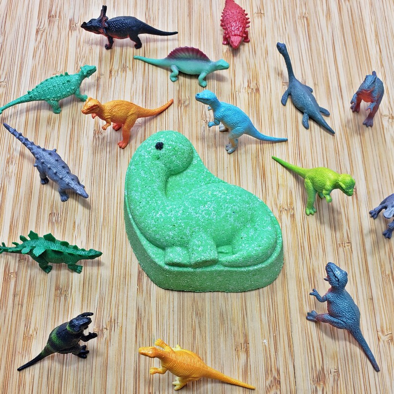 May include: A green bath bomb shaped like a dinosaur, surrounded by a variety of colorful plastic dinosaur toys on a wooden surface.