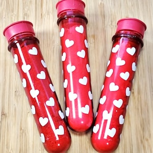 May include: Three red tubes with white heart patterns and pink caps. The tubes are filled with a red substance. The tubes are arranged on a wooden surface.