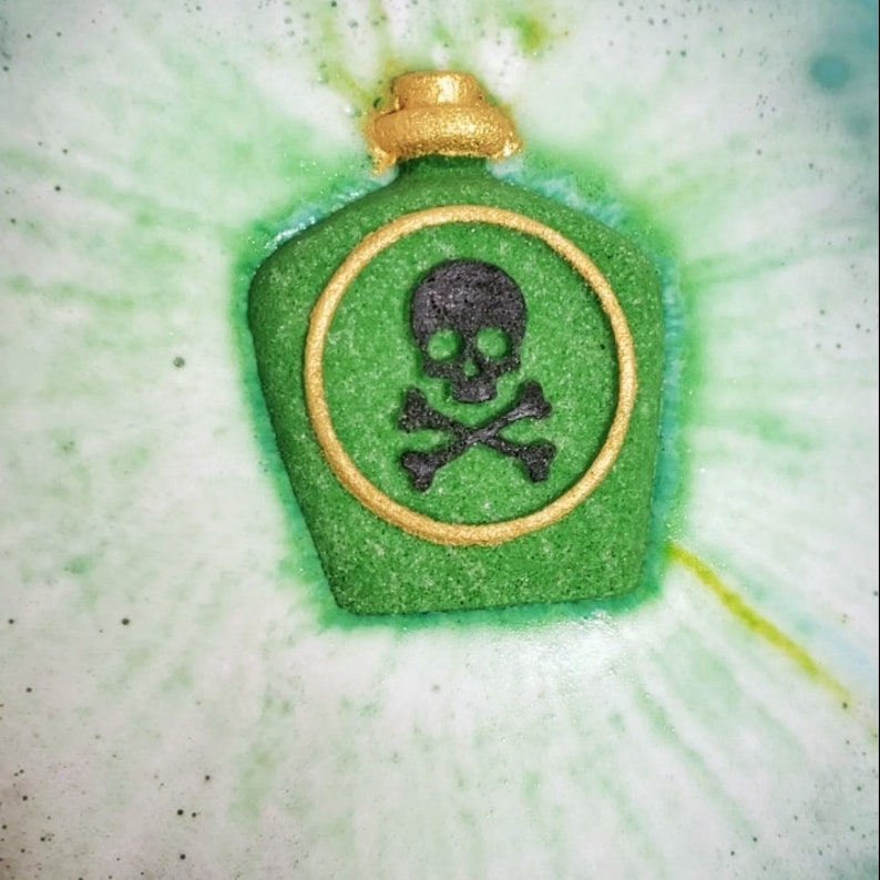 May include: A green bath bomb shaped like a poison bottle, with a gold-colored top and a black skull and crossbones emblem. The bath bomb is surrounded by green and white water, creating a bubbly effect.