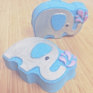 May include: Two blue bath bombs shaped like elephants with silver glitter accents. Each elephant has a pink flower design coming out of its trunk.