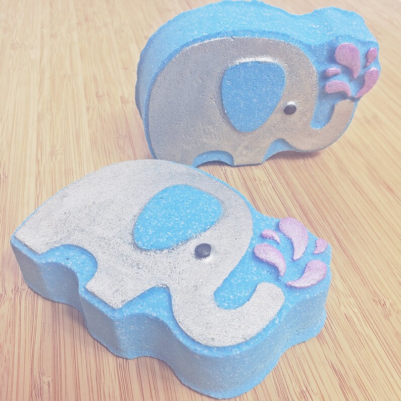 May include: Two blue bath bombs shaped like elephants with silver glitter accents. Each elephant has a pink flower design coming out of its trunk.