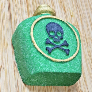 May include: A green, bottle-shaped bath bomb with a gold-colored top and a gold-lined circle featuring a black skull and crossbones. The bath bomb has a textured surface and is designed to resemble a poison bottle.