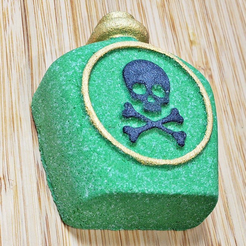 May include: A green, bottle-shaped bath bomb with a gold-colored top and a gold-lined circle featuring a black skull and crossbones. The bath bomb has a textured surface and is designed to resemble a poison bottle.