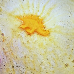 May include: A yellow dinosaur bath bomb floating in a white and yellow bubbly bath.