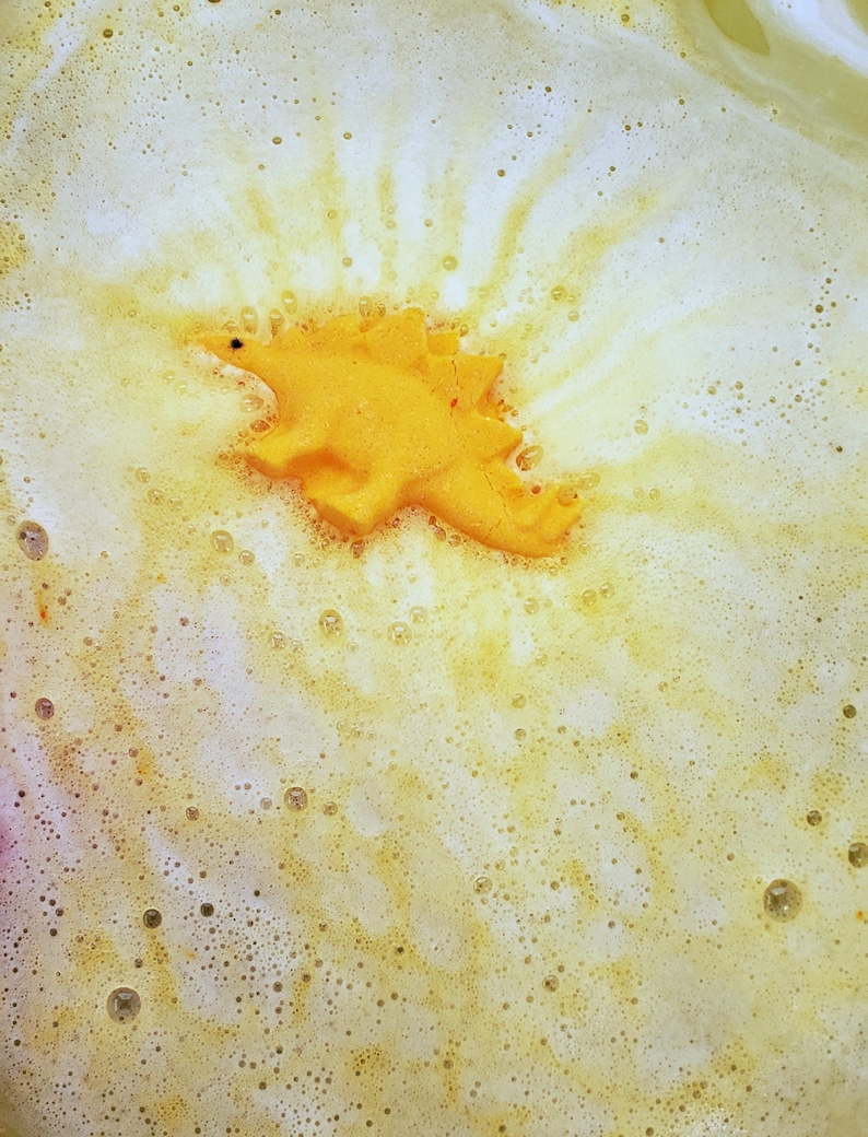 May include: A yellow dinosaur bath bomb floating in a white and yellow bubbly bath.