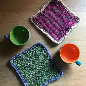 以下が含まれることがあります： 木製の表面に、緑と青の鍋つかみと、ピンクと白の鍋つかみの2つが置かれています。鍋つかみの隣には、空のマグカップが2つあります。