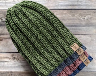 May include: Four handmade crochet beanies in green, blue, and pink. The beanies are made with a ribbed stitch and have small tags attached. The beanies are displayed on a wooden surface.