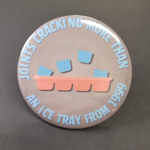 May include: A round button with the text "JOINTS CRACKING MORE THAN AN ICE TRAY FROM 1999" in light blue. The button has a gray background with a pink ice tray and blue ice cubes.
