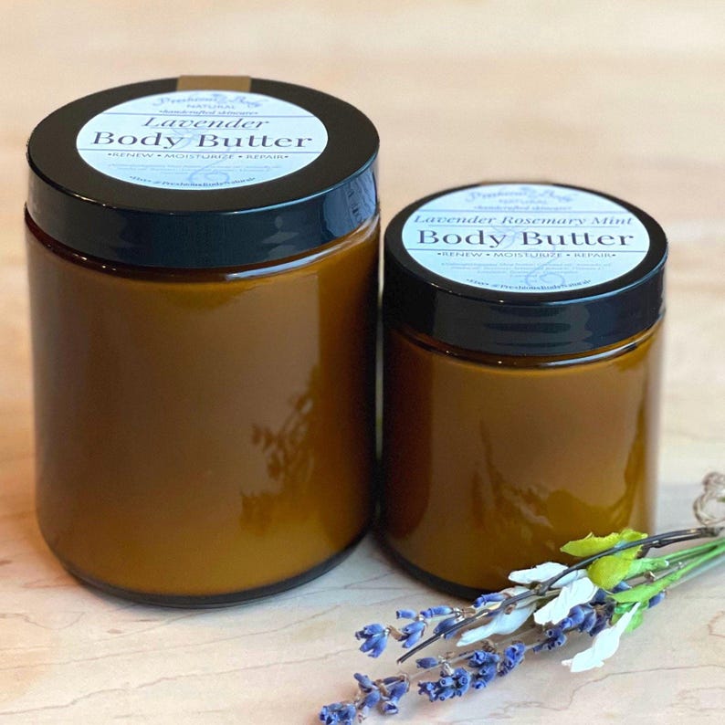 To Amber glass jars of body butter with black lids and white labels sitting on a wooden surface
