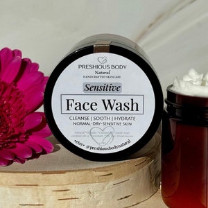 May include: A brown jar with a white label that reads 'PRESHIOUS BODY Natural HANDCRAFTED SKINCARE Sensitive Face Wash CLEANSE | SOOTH | HYDRATE NORMAL-DRY-SENSITIVE SKIN Natural Organic-Coconut Oil Castile soap essential oils of Lavender, *yer, Frankincense etsy @preshiousbodynatural' The jar is sitting on a wooden surface with a pink flower and a second jar of face wash in the background.