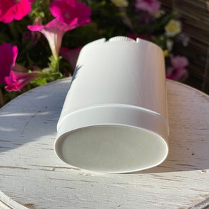 May include: A white deodorant container with a light gray deodorant stick inside. The container is cylindrical with a slightly domed top and a smooth, flat bottom. The deodorant stick is visible through the transparent bottom of the container. The container is shown on a white wooden surface.