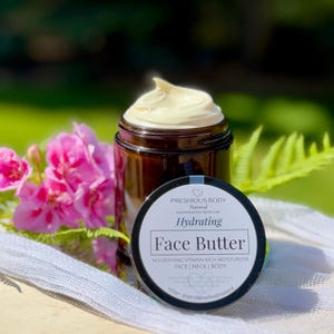 May include: A glass jar with a black lid filled with a face butter. The jar has a white label with the text 'PRESHIOUS BODY', 'Natural', 'HANDCRAFTED SKINCARE', 'Hydrating', 'Face Butter', 'NOURISHING VITAMIN RICH MOISTURIZER', 'FACE | NECK | BODY', 'Natural Organic Shea butter Rosehip oil Coconut Vitamin E Essential oils of Frankincense Lavender', 'etsy. @preshiousbodynatural'.