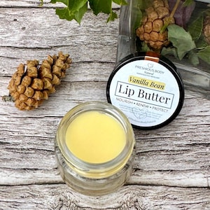 May include: A small jar of yellow lip butter with a black lid. The lid has the text "Precious Body Natural Handcrafted Naturals Vanilla Bean Lip Butter Nourish Renew Protect" on it.