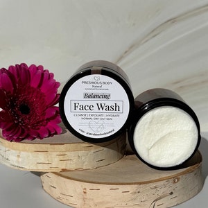 Puede incluir: Un tarro negro con una etiqueta blanca que dice "PRESHIOUS BODY Natural HANDCRAFTED SKINCARE Balancing Face Wash CLEANSE | EXFOLIATE | HYDRATE NORMAL-DRY-OILY SKIN elsy @preshiousbodynatural". El tarro est&aacute; sobre una superficie de madera con una flor rosa en el fondo.