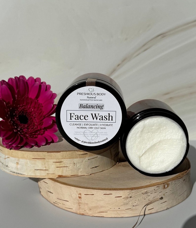 Puede incluir: Un tarro negro con una etiqueta blanca que dice "PRESHIOUS BODY Natural HANDCRAFTED SKINCARE Balancing Face Wash CLEANSE | EXFOLIATE | HYDRATE NORMAL-DRY-OILY SKIN elsy @preshiousbodynatural". El tarro est&aacute; sobre una superficie de madera con una flor rosa en el fondo.