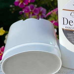 Close-up view of a white deodorant container with a light gray deodorant stick inside. The container is cylindrical with a slightly oval-shaped base.  The label on the adjacent container reads 'Clay Deodorant' and 'MAGNESIUM'.  Ingredients listed include coconut oil and bentonite clay. The background is blurred, showing purple flowers.