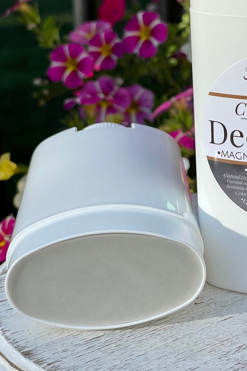 Close-up view of a white deodorant container with a light gray deodorant stick inside. The container is cylindrical with a slightly oval-shaped base.  The label on the adjacent container reads 'Clay Deodorant' and 'MAGNESIUM'.  Ingredients listed include coconut oil and bentonite clay. The background is blurred, showing purple flowers.