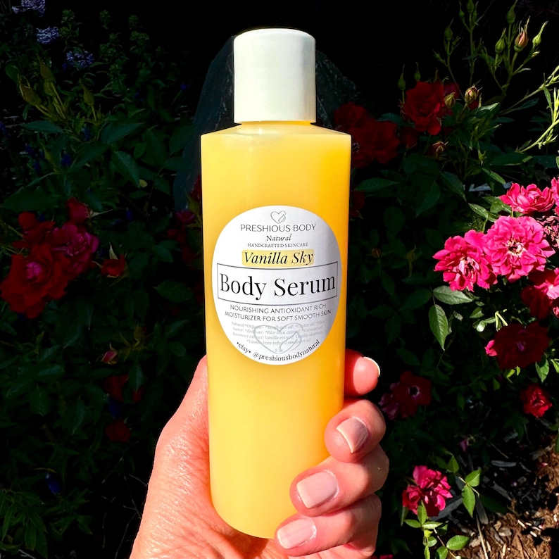 A bottle of body serum with a white label. A hand is holding the bottle. The label says 'Preshious Body Natural Handcrafted Skincare’ Body Serum Nourishing Antioxidant Rich Moisturizer for Soft Smooth Skin *etsy@preshiousbodynatural'