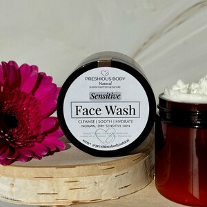May include: A brown jar with a white label that reads 'PRESHIOUS BODY Natural HANDCRAFTED SKINCARE Sensitive Face Wash CLEANSE | SOOTH | HYDRATE NORMAL-DRY-SENSITIVE SKIN Natural *Organic* Coconut Castile soap essential oil of Lavender, Tea Tree, Frankincense *etsy* @preshiousbodynatural' The jar is sitting on a wooden surface next to a clear jar with a white lid and white cream inside.