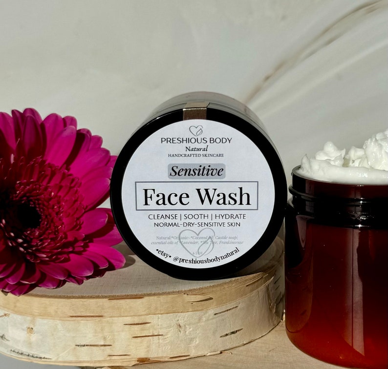 May include: A brown jar with a white label that reads 'PRESHIOUS BODY Natural HANDCRAFTED SKINCARE Sensitive Face Wash CLEANSE | SOOTH | HYDRATE NORMAL-DRY-SENSITIVE SKIN Natural *Organic* Coconut Castile soap essential oil of Lavender, Tea Tree, Frankincense *etsy* @preshiousbodynatural' The jar is sitting on a wooden surface next to a clear jar with a white lid and white cream inside.