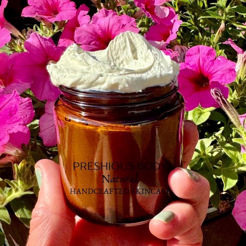 amber glass jar filled with fluffy and creamy white body butter. There is a hand holding the jar and pink flowers in the background.