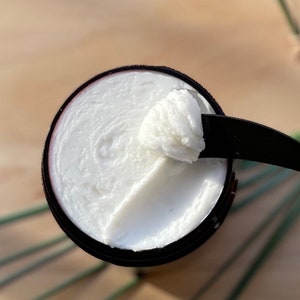 May include: A close-up of a white cream in a brown container. The cream is thick and has a smooth texture. A small amount of cream has been scooped out of the container with a black spatula.