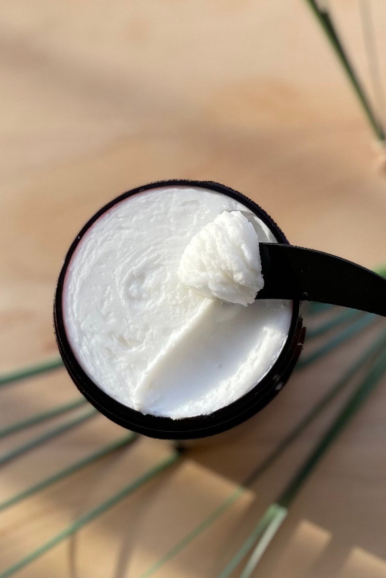 May include: A close-up of a white cream in a brown container. The cream is thick and has a smooth texture. A small amount of cream has been scooped out of the container with a black spatula.