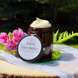 May include: A glass jar with a black lid filled with a face butter. The jar has a white label with the text 'PRESHIOUS BODY', 'Natural', 'HANDCRAFTED SKINCARE', 'Hydrating', 'Face Butter', 'NOURISHING VITAMIN RICH MOISTURIZER', 'FACE | NECK | BODY', 'Natural Organic Shea butter Rosehip oil Coconut Vitamin E Essential oils of Frankincense Lavender', 'etsy. @preshiousbodynatural'.