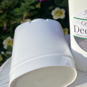 May include: A white deodorant stick with a label that reads 'Cedarwood Deodorant' and 'MAGNESIUM HYDROXIDE'. The deodorant is sitting on a white wooden surface.