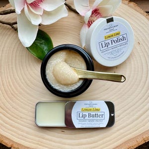 Puede incluir: Un conjunto de dos productos para el cuidado de los labios con aroma a lima limón. El exfoliante labial está en un frasco de vidrio con una cuchara dorada. La manteca labial está en una pequeña lata de metal. Ambos productos están sobre una superficie de madera con flores blancas.