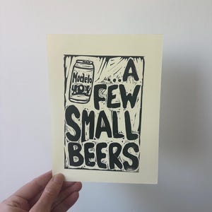 Puede incluir: Una impresión de color crema con texto negro y una ilustración de una lata de cerveza Modelo. El texto dice "A FEW SMALL BEERS". La impresión está sostenida por una mano contra una pared blanca.