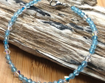 May include: A delicate bracelet featuring light blue beads and iridescent crystal accents. The bracelet is secured with a silver-tone clasp. The beads are approximately 0.5 cm in diameter. The bracelet is displayed on a piece of driftwood.