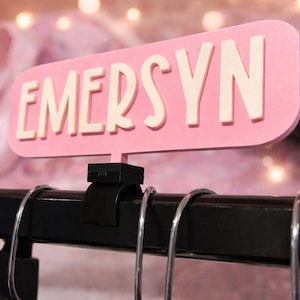 May include: A pink sign with the name "EMERSYN" in white letters is attached to a black metal bar. The sign is rectangular with rounded corners. The background is blurred, with pink ballet shoes and a pink fluffy item.