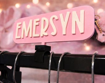 May include: A pink sign displaying the name "EMERSYN" in white lettering. The sign is mounted on a black metal bar with silver rings. The background is blurred, featuring pink ballet shoes and a fluffy pink accessory.