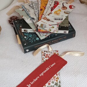 May include: A set of handmade fabric bookmarks with various designs, including musical instruments, cars, and floral patterns. The bookmarks have a red ribbon attached to them. One bookmark has the text "La lecture agrandit l'âme" embroidered on it.