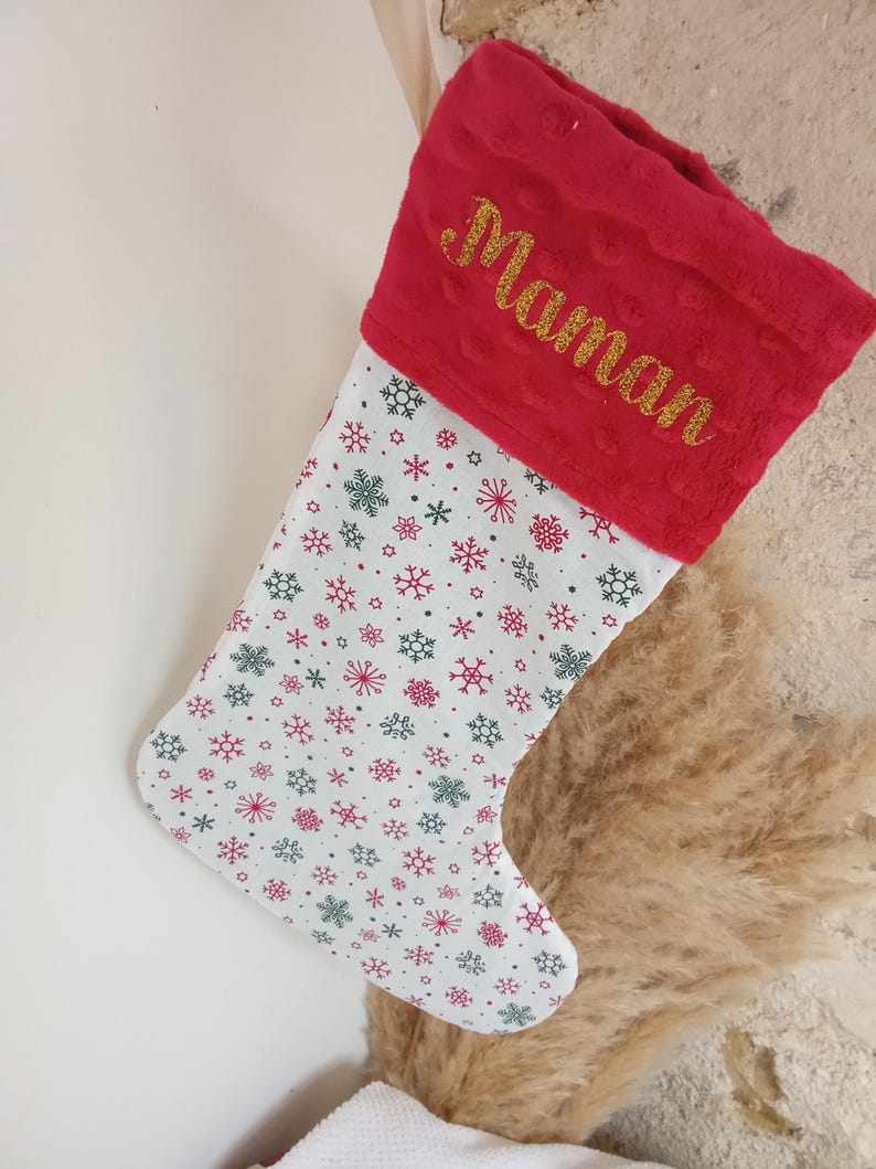 K&ouml;nnte beinhalten: Roter und wei&szlig;er Weihnachtssocken mit Schneeflockenmuster und dem Wort "Maman" in goldenen Glitzer auf dem B&uuml;ndchen gestickt.