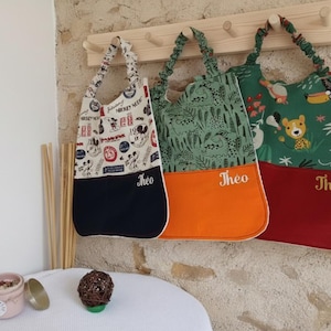 Peut inclure: Trois bavoirs en tissu avec des tours de cou élastiqués froncés, chacun portant le prénom Théo brodé en doré. Le premier bavoir présente un motif Mickey Mouse sur fond blanc et une base bleu marine, le deuxième un motif dinosaure vert et une base orange, et le troisième un motif animaux de la jungle et une base rouge.