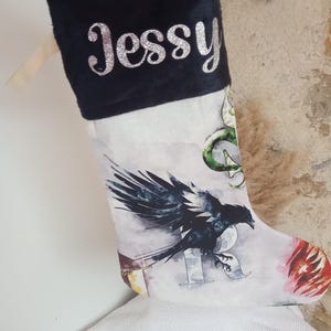 Peut inclure: Une chaussette de Noël blanche avec un revers noir et le nom "Jessy" en paillettes argentées. La chaussette présente un motif aquarelle d'un corbeau, d'un blason de la maison de Poudlard et d'un dragon.